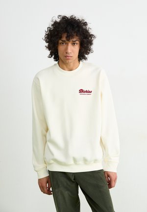 Crème-kleurige sweatshirt met een ronde hals, ribbelboorden en onderkant; heeft een geborduurd bordeauxrood logo op de borst. Zachte stoftextuur.