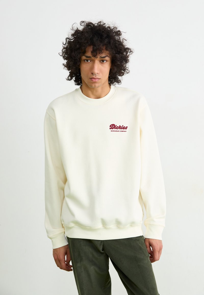 Cream-colored sweatshirt with a round neck, ribbed cuffs, and hem; features a embroidered maroon logo on the chest. Soft fabric texture.