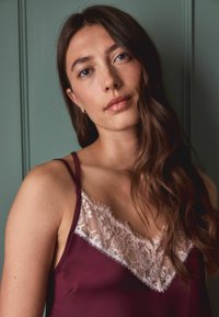 Burgunder Satin-Camisole mit einem Spitzenrand am Ausschnitt. Verfügt über dünne Träger und eine glatte Textur, vor einem grünen Hintergrund.