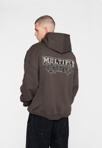Felpa oversize marrone scuro con tasca frontale, cappuccio e testo grafico "MULTIPLY APPAREL" ben visibile sulla parte posteriore.