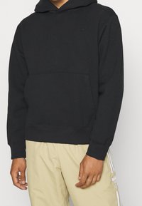 Svart huvtröja i bomullsblandning med framficka och ribbade ärmar. Har en liten, ton-i-ton logotyp på bröstet.