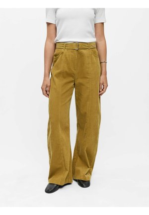 Femme portant un pantalon large en velours côtelé jaune moutarde avec ceinture, un haut blanc à manches courtes, des chaussures noires et un bracelet argenté au poignet droit.