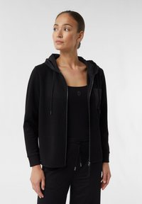 Schwarze Zip-Hoodie mit einer Kordelzugkapuze und subtiler Vorderasche. Der Stoff ist glatt und das Design weist eine taillierte Silhouette auf.