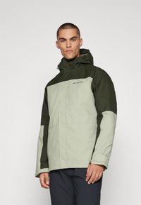 Veste imperméable vert clair et vert foncé avec capuche, fermeture éclair et poignets ajustables ; comporte le logo Columbia sur la poitrine.