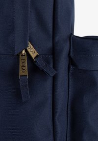 Tmavě modrá látka batohu s texturovaným povrchem, doplněná zlatými zipy a kováním, s logovanými jezky pro zvýraznění detailů.