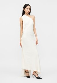 Pikkade tumedate juustega naine, kes kannab valget üheõlalist maani ulatuvat kleiti ja musti teravate ninadega kontsakingi, millel on pahkluune rihmad.