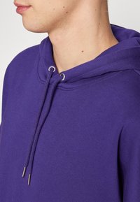 Felpa viola con cappuccio e coulisse, caratterizzata da una texture morbida e scollo arrotondato. Dettagli metallici alle estremità delle coulisse.