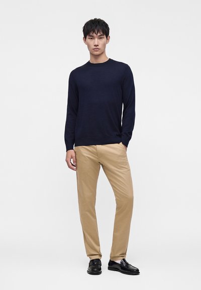 Mand iført en mørk marineblå sweater, beige bukser og sorte finere sko, der står mod en ensfarvet hvid baggrund.