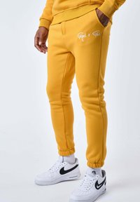 Los pantalones jogger de algodón amarillo presentan un diseño ajustado con puños elásticos y bolsillos laterales. El logo de la marca está bordado en blanco al frente.