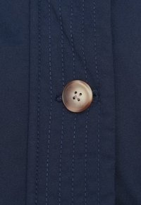 Bottone marrone a quattro fori cucito su tessuto blu scuro con linee di cucitura verticali.