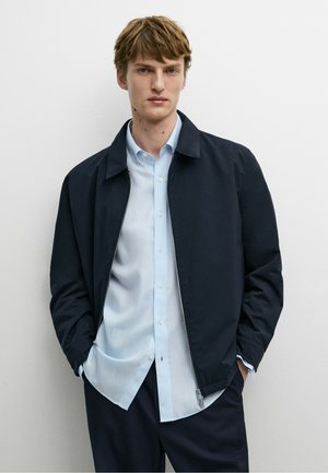 Ung mand med kort lyst brunt hår iført en lyseblå skjorte med knapper og en mørk navy lynlåsjakke, stående mod en almindelig hvid væg.