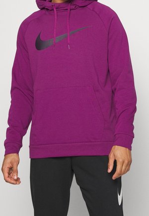 Mężczyzna w fioletowej bluzie z kapturem Nike z czarnym logo Swoosh i czarnych spodniach z białym logo Nike Swoosh, stojący na jednolitym tle.