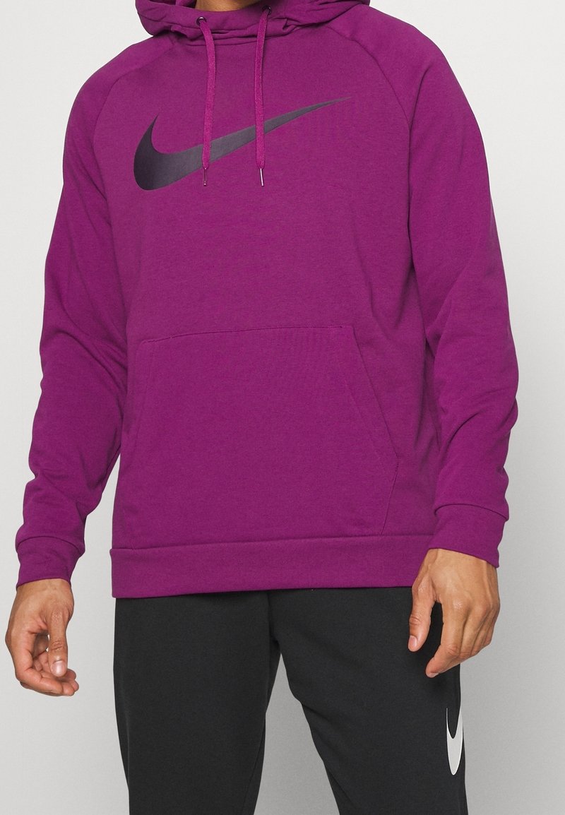 Homme portant un sweat à capuche violet Nike avec le logo swoosh noir et un pantalon noir avec le logo swoosh blanc Nike, debout devant un fond uni.