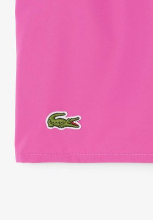 Rosa Baumwollstoff mit glatter Textur, versehen mit einem gestickten grünen Krokodil-Logo in der unteren linken Ecke.