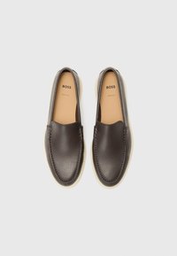 Paire de mocassins en cuir marron foncé sans lacets avec dessus texturé et semelles intérieures beiges portant l’étiquette "BOSS" sur un fond clair uni.