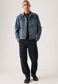 Τζιν μπουφάν σε ανοιχτό μπλε χρώμα με εμπρός τσέπες, συνδυασμένο με σκουρόχρωμο παντελόνι καρό με πλαϊνές τσέπες και μαύρα αθλητικά παπούτσια.