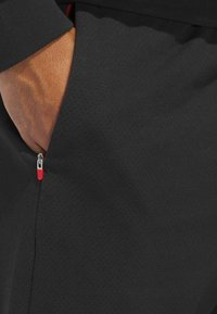 Zwarte sportshorts van een textuur en geperforeerde stof. Voorzien van een rits met rode accenten op de linkerzak.