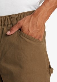 Brune bomuldsbukser har en sidelomme med synlig syning. Den elastiske talje sørger for en sikker pasform. Fingeren hviler i lommen.