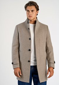 Mand iført en lysbrun uldfrakke med sorte knapper over en hvid skjorte og blå jeans, stående foran en ensfarvet baggrund.