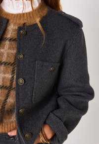 Veste en mélange de laine grise avec des boutons en ton doré, dotée d'une poche poitrine et d'épaules structurées. Portée sur un pull à carreaux marron.