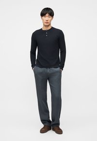 Άνδρας που στέκεται με τα χέρια στις τσέπες, φοράει μαύρο μακρυμάνικο πουκάμισο henley, γκρι παντελόνι και καφέ αθλητικά παπούτσια σε απλό φόντο.