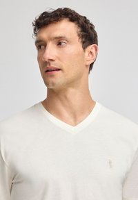 T-shirt crema a scollo a V realizzato in tessuto morbido, caratterizzato da un sottile motivo testurizzato e un piccolo logo ricamato sul lato sinistro del petto.