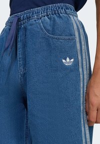 Pantaloni jogger in denim blu con elastico in vita, caratterizzati da un cordino viola, tasche laterali e un logo ricamato bianco sulla gamba sinistra.