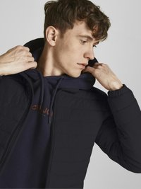 Giacca a vento nera con finitura testurizzata, dotata di cappuccio e chiusura con zip. Indossata sopra una felpa blu con un logo a contrasto.