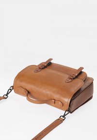 Sac bandoulière en cuir marron clair avec deux fermetures à rabat à boucle, une petite poignée et une bandoulière réglable sur fond blanc.
