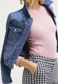 Denim jacket with a collar, light blue color, two chest pockets, and copper buttons, paired with a ribbed pink top and houndstooth-patterned pants.