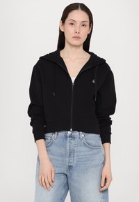 Crna skraćena hoodie majica s patentnim zatvaračem, podesivim vezicama i rebrastim manžetama. Uparena s svijetloplavim trapericama visokog struka.