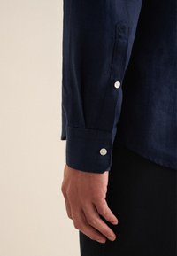 Marineblauwe linnen blouse met een knoopmanchet met witte knopen, zichtbare stiksels en een subtiele geborduurde detail op de mouw.