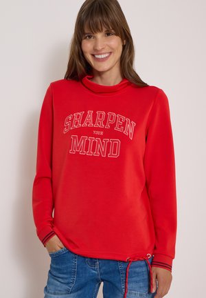 Femme souriante portant un sweat-shirt rouge avec le texte "SHARPEN YOUR MIND" et un jean bleu, debout devant un fond clair uni.