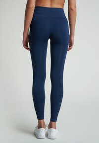 Námořnické modré legíny s vysokým pasem a hladkou texturou. Mají přiléhavý střih a jemné postranní švy. Nosené s bílými teniskami.