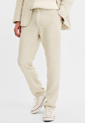 Pantalons en lin beige à coupe droite, dotés de deux poches avant, avec une texture subtile, associés à des baskets blanches.