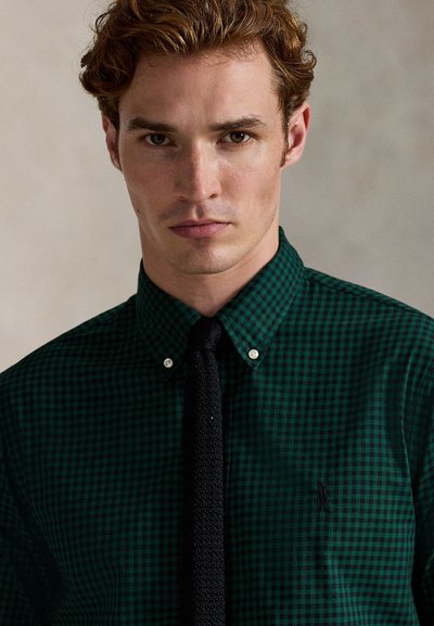 Young man with light brown hair wearing a dark green checkered shirt and black knit tie, looking straight ahead with a neutral expression.
