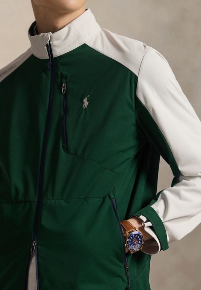 Personne portant une veste zippée verte et blanche avec une poche poitrine et une montre au cadran bleu avec un bracelet en cuir marron au poignet gauche.