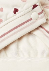 Tessuto chiaro con piccoli motivi a forma di cuore in rosa e bordeaux. Presenta una banda a righe con un bottone a pressione bianco per la chiusura.