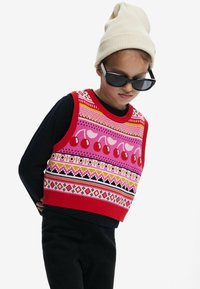 Maglione senza maniche lavorato a maglia con un motivo rosso, rosa e nero con ciliegie e design geometrici; abbinato a una maglietta nera a maniche lunghe e a un berretto beige.