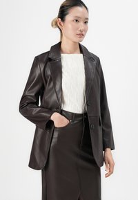 Vrouw in een donkerbruin leren blazer en rok met een witte textuurtop, staand tegen een effen witte achtergrond.