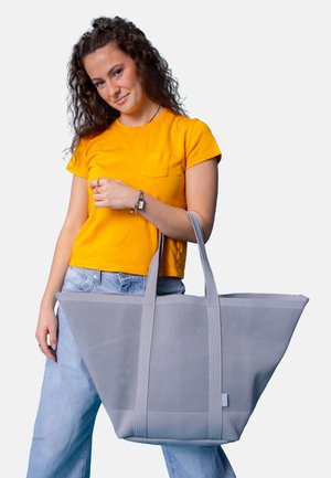 Woman in a yellow shirt and jeans holding a large gray mesh tote bag by its handles, standing against a plain background.