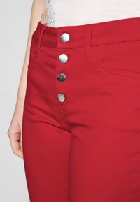 Pantalon ajusté rouge avec une texture lisse, doté d'une fermeture à bouton avec quatre boutons métalliques et d'une poche latérale, partiellement visible.
