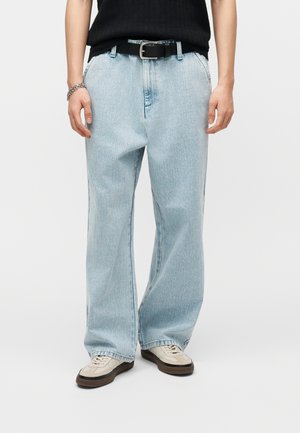 Persona con vaqueros anchos de pierna en azul claro, cinturón negro, top sin mangas negro, zapatillas beige y una pulsera plateada sobre un fondo liso.