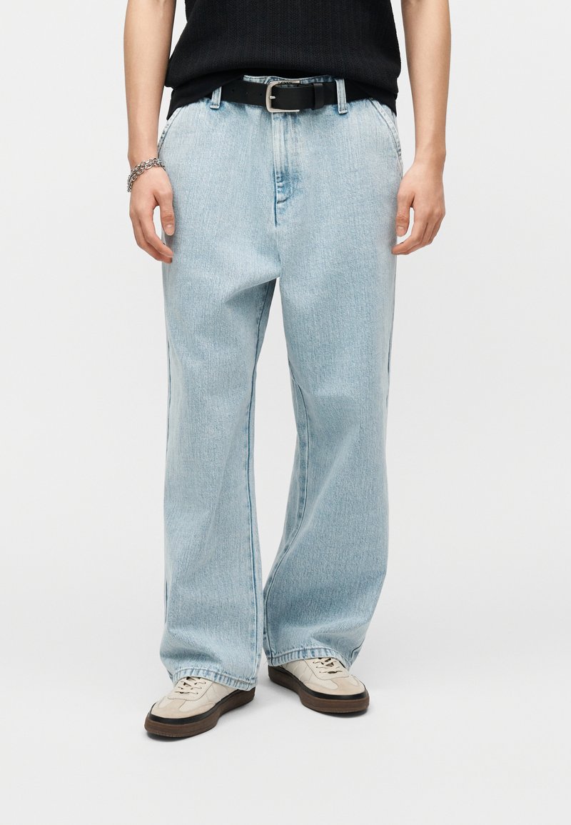 Άτομο που φοράει ανοιχτό μπλε τζιν παντελόνι με φαρδιά μπότα, μαύρη ζώνη, μαύρο αμάνικο τοπ, μπεζ αθλητικά παπούτσια και ασημένιο βραχιόλι μπροστά σε απλό φόντο.