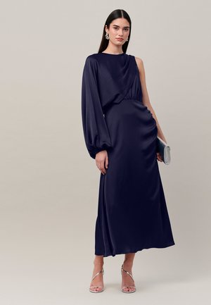 Kvinde iført en lang navy asymmetrisk kjole med én lang ærme, der holder en sølv clutch og bærer sølv hæle og blomsterøreringe.
