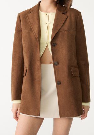 Vrouw draagt een bruine suède blazer over een crème cropped gebreide cardigan met knopen en een beige minirok tegen een effen achtergrond.