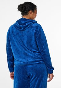 Blauer Samt-Kapuzenpullover mit Sternmuster. Weiche Textur mit Kapuze und gerippten Bündchen. Sweatpants im passenden Design vervollständigen das Outfit.