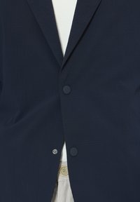 Marineblazer aus strukturiertem Stoff, mit zwei Knöpfen, einem Revers mit Kerbung und einem dezenten Steppmuster. Darunter ist ein helles Hemd zu sehen.