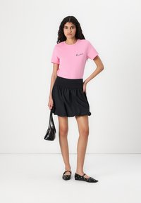 T-shirt en coton rose avec texte brodé noir, associé à une jupe noire à taille élastique et à la coupe volumineuse. Sac à main et chaussures noires.