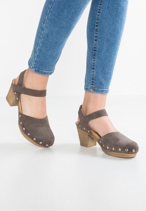 Brązowe zamszowe sandały na obcasie z zaokrąglonym noskiem, paskiem wokół kostki i ozdobnymi srebrnymi ćwiekami wzdłuż krawędzi; drewniany obcas bloczkowy.